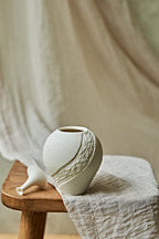 A small matte white ceramic porcelain oil lamp with a textured diagonal wave design, displayed on a rustic wooden stool draped with a neutral linen cloth.

