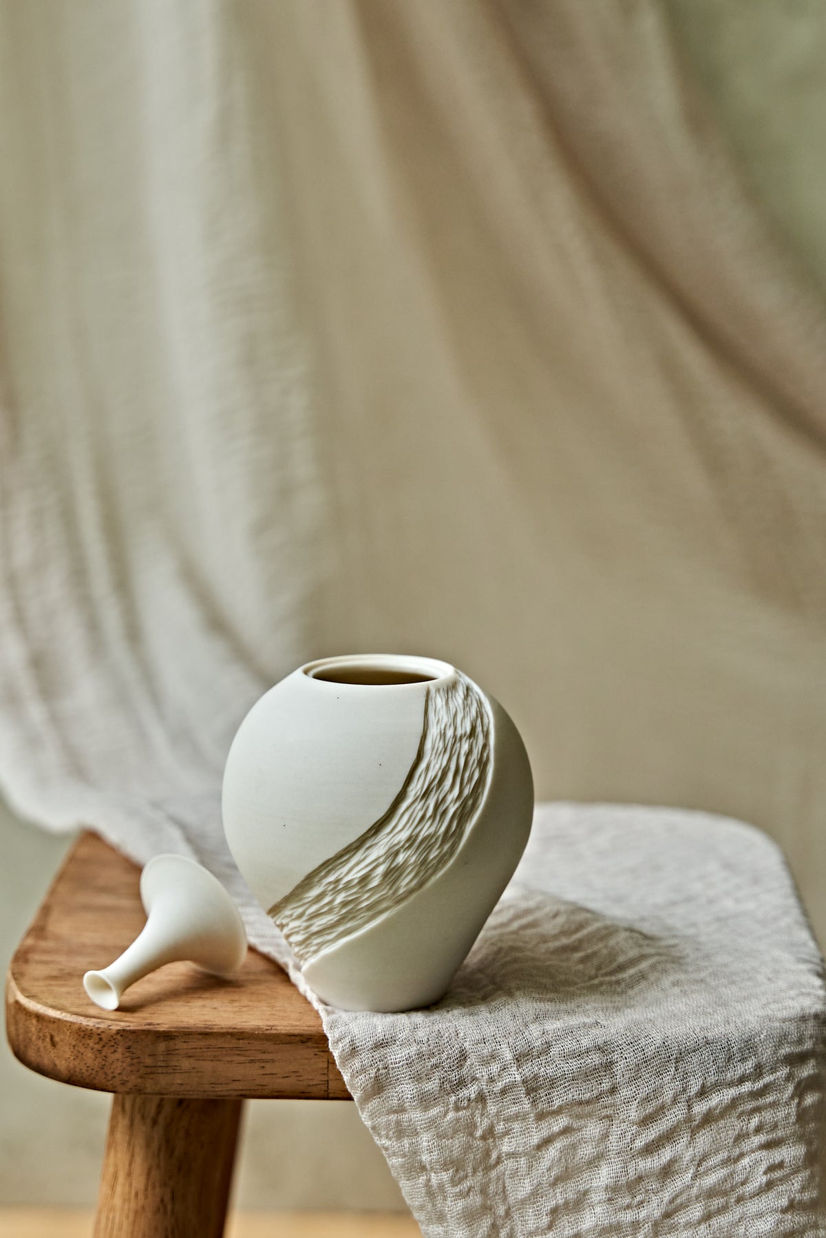 A small matte white ceramic porcelain oil lamp with a textured diagonal wave design, displayed on a rustic wooden stool draped with a neutral linen cloth.

