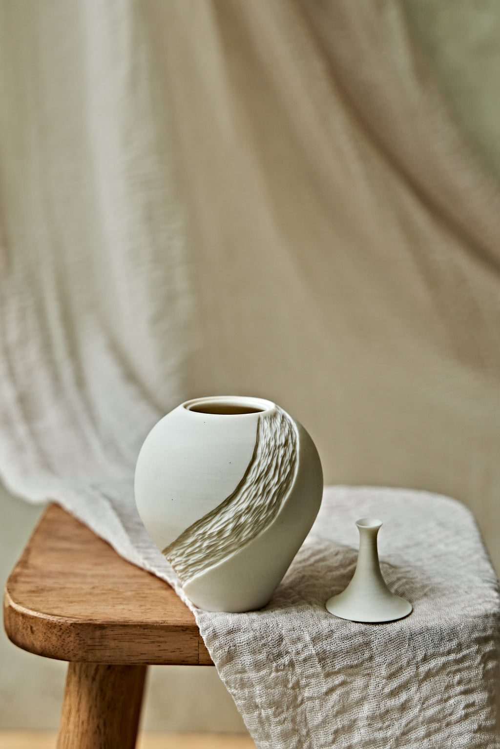 A small matte white ceramic porcelain oil lamp with a textured diagonal wave design, displayed on a rustic wooden stool draped with a neutral linen cloth.
