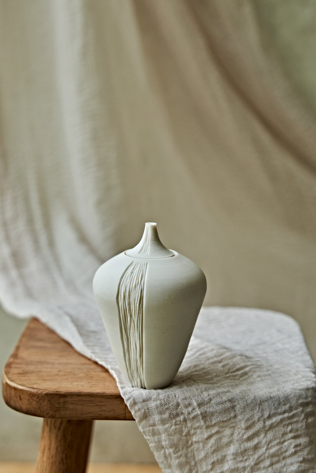 A small matte ceramic oil lamp with a narrow neck and vertical textured lines, displayed on a wooden stool draped with light beige linen fabric.

