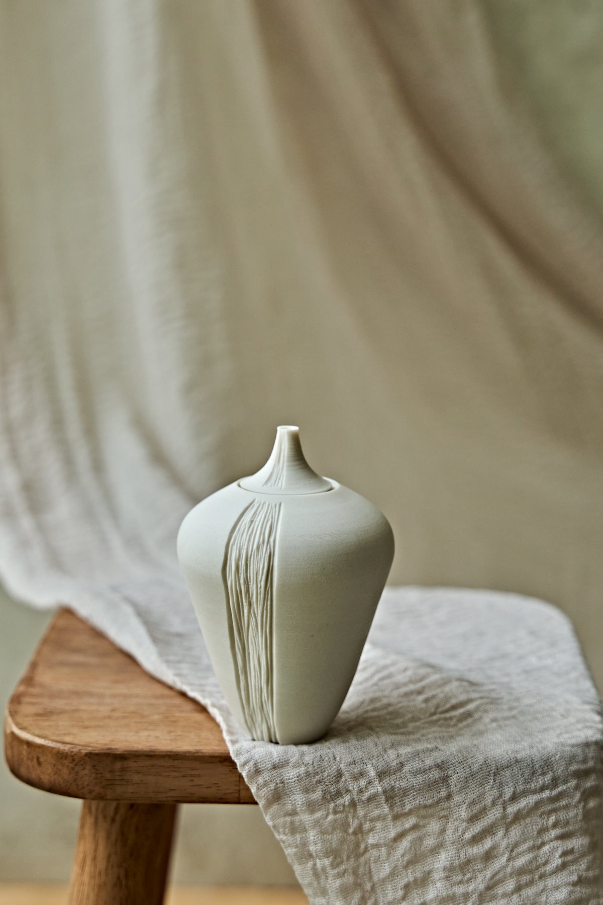 A small matte ceramic oil lamp with a narrow neck and vertical textured lines, displayed on a wooden stool draped with light beige linen fabric.

