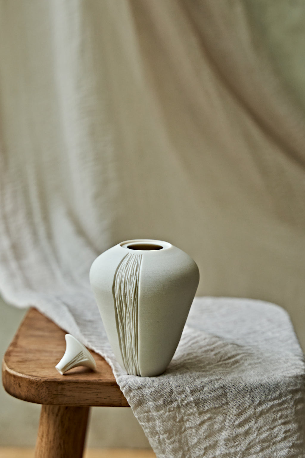 A small matte ceramic oil lamp with a narrow neck and vertical textured lines, displayed on a wooden stool draped with light beige linen fabric.