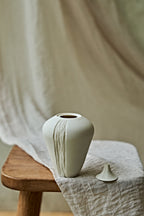A small matte ceramic oil lamp with a narrow neck and vertical textured lines, displayed on a wooden stool draped with light beige linen fabric.

