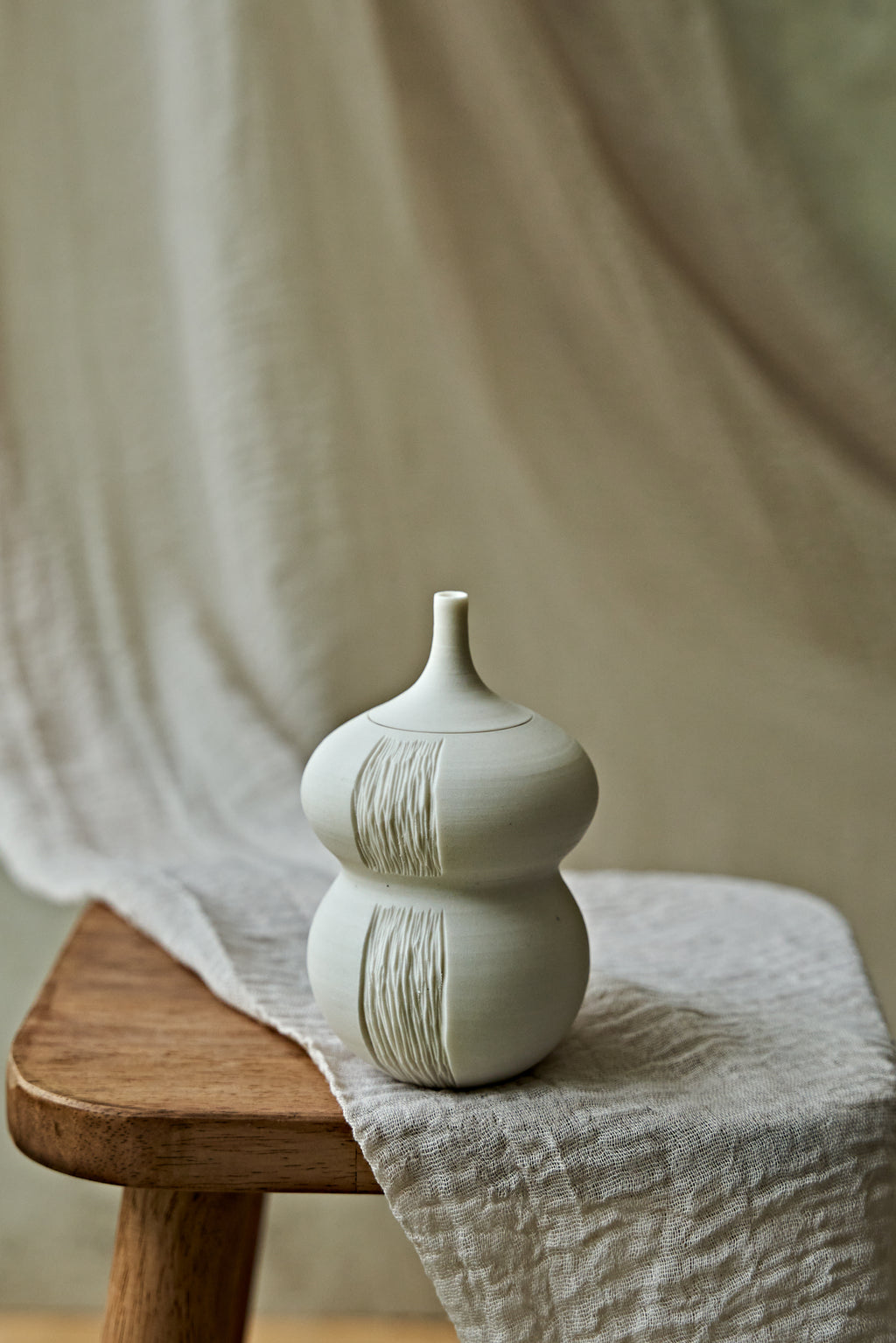 A small matte ceramic oil lamp with a rounded double-bulb shape and vertical textured ridges, displayed on a wooden stool draped with a light fabric. A small matching ceramic object rests beside it against a soft neutral backdrop.

