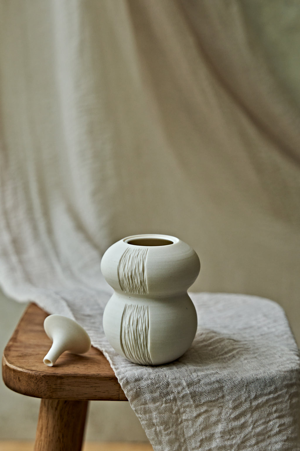 A small matte ceramic oil lamp with a rounded double-bulb shape and vertical textured ridges, displayed on a wooden stool draped with a light fabric. A small matching ceramic object rests beside it against a soft neutral backdrop.

