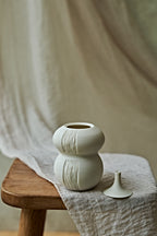 A small matte ceramic oil lamp with a rounded double-bulb shape and vertical textured ridges, displayed on a wooden stool draped with a light fabric. A small matching ceramic object rests beside it against a soft neutral backdrop.