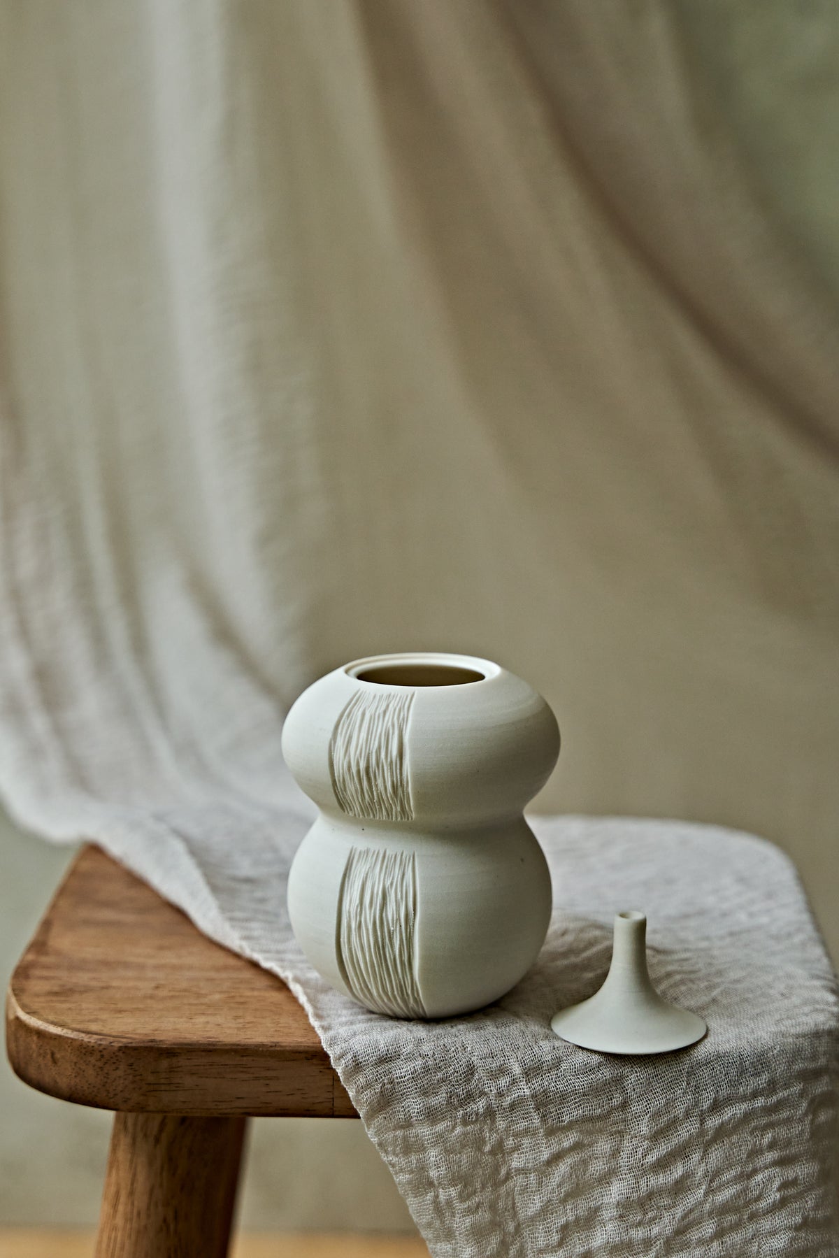 A small matte ceramic oil lamp with a rounded double-bulb shape and vertical textured ridges, displayed on a wooden stool draped with a light fabric. A small matching ceramic object rests beside it against a soft neutral backdrop.