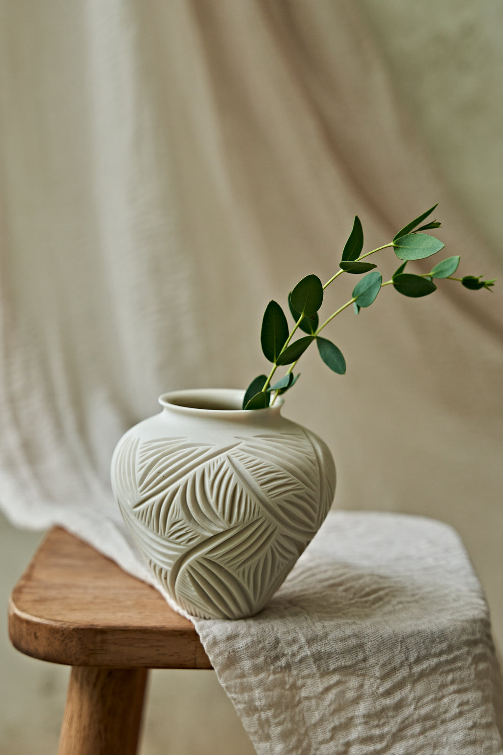 A small off-white ceramic vase with a carved leaf pattern sits on a wooden stool draped with a light fabric, holding a green leafy sprig against a soft neutral background.