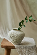 A small off-white ceramic vase with a carved leaf pattern sits on a wooden stool draped with a light fabric, holding a green leafy sprig against a soft neutral background.