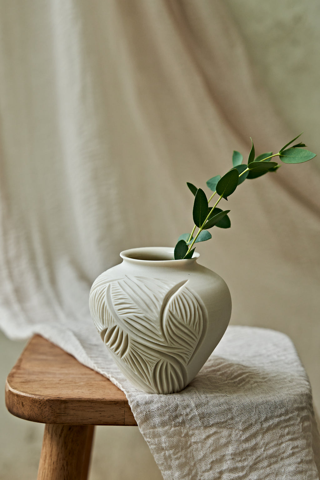 A small off-white ceramic vase with a carved leaf pattern sits on a wooden stool draped with a light linen cloth, holding a green eucalyptus branch against a soft neutral fabric backdrop.