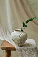 A small off-white ceramic vase with a carved leaf pattern sits on a wooden stool draped with a light linen cloth, holding a green eucalyptus branch against a soft neutral fabric backdrop.