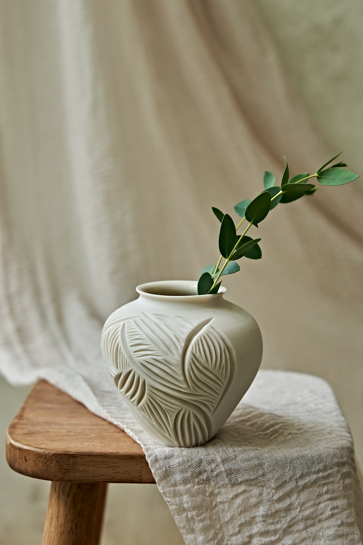A small off-white ceramic vase with a carved leaf pattern sits on a wooden stool draped with a light linen cloth, holding a green eucalyptus branch against a soft neutral fabric backdrop.