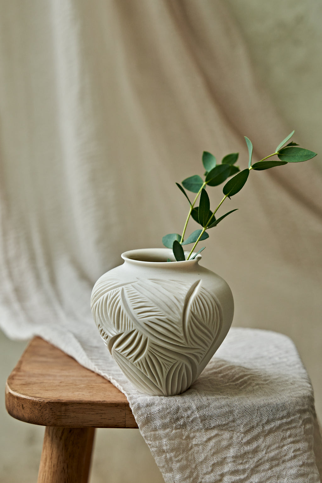 A small ceramic vase with a matte off-white finish and embossed leaf patterns, holding a green eucalyptus sprig, styled on a wooden stool draped with a neutral linen cloth.