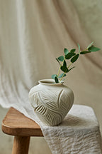 A small ceramic vase with a matte off-white finish and embossed leaf patterns, holding a green eucalyptus sprig, styled on a wooden stool draped with a neutral linen cloth.