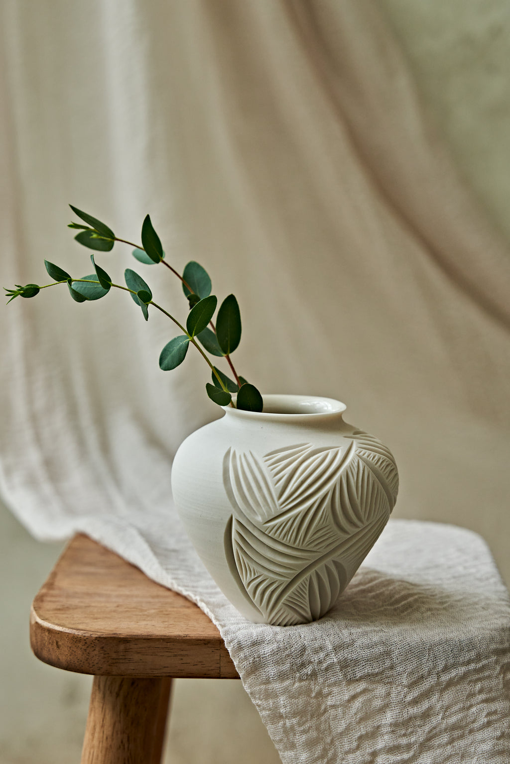 A small white ceramic vase with embossed leaf patterns sits on a wooden stool draped with a cream linen cloth, holding a green eucalyptus sprig against a soft neutral fabric backdrop.