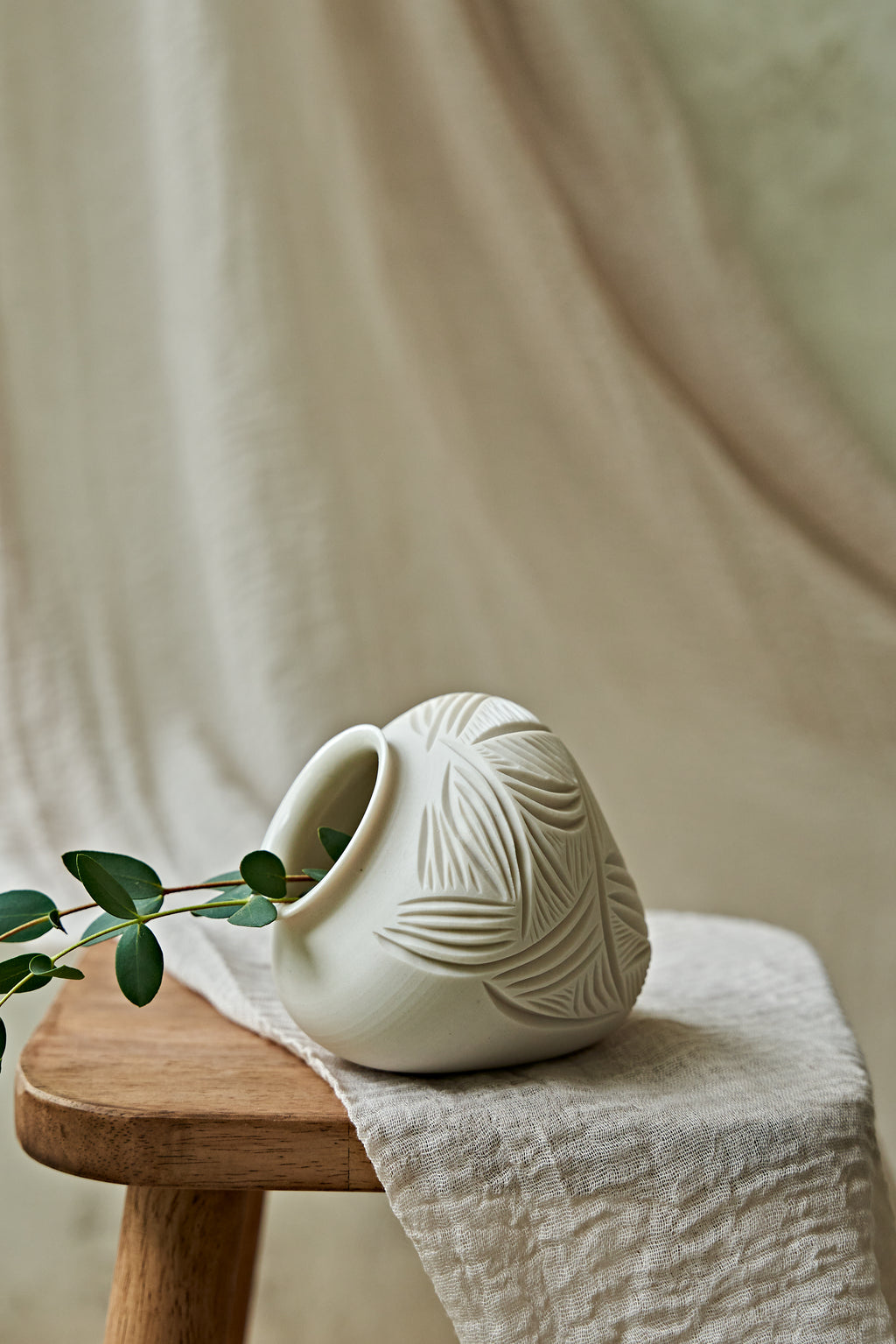 A small off-white ceramic pitcher with a leaf embossed pattern, resting on a wooden stool draped with a linen cloth, with a green leafy sprig in its opening.