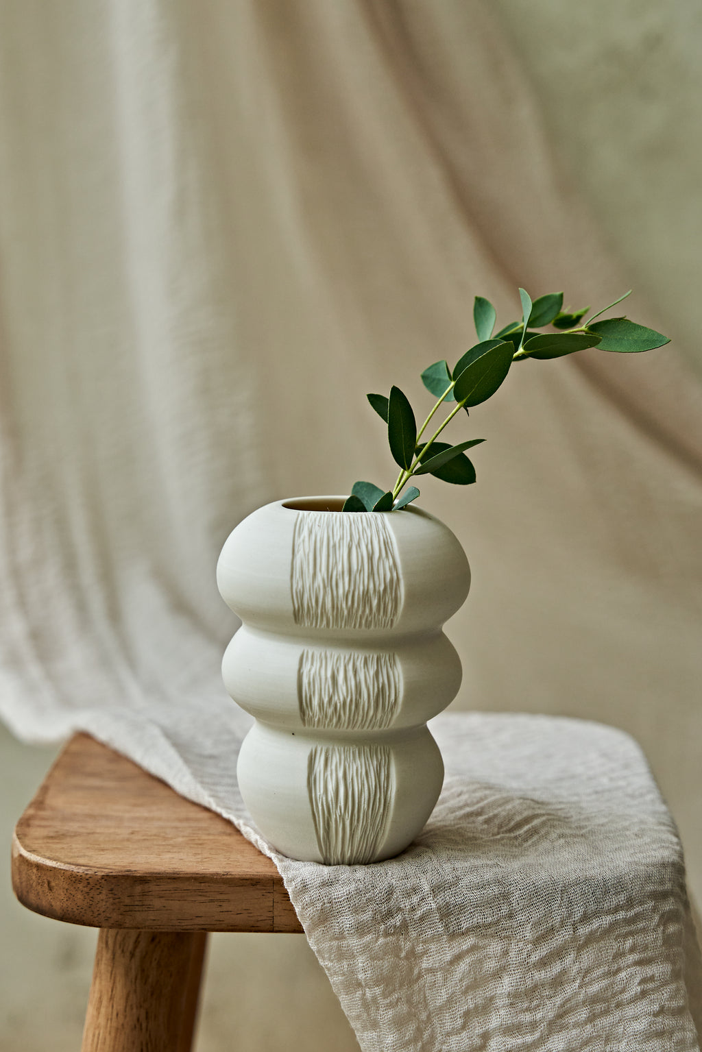 A textured, off-white ceramic vase with a stacked, rounded design sits on a wooden stool draped with a neutral linen cloth. A small green leafy sprig is placed inside the vase.
