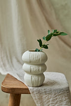 A textured, off-white ceramic vase with a stacked, rounded design sits on a wooden stool draped with a neutral linen cloth. A small green leafy sprig is placed inside the vase.