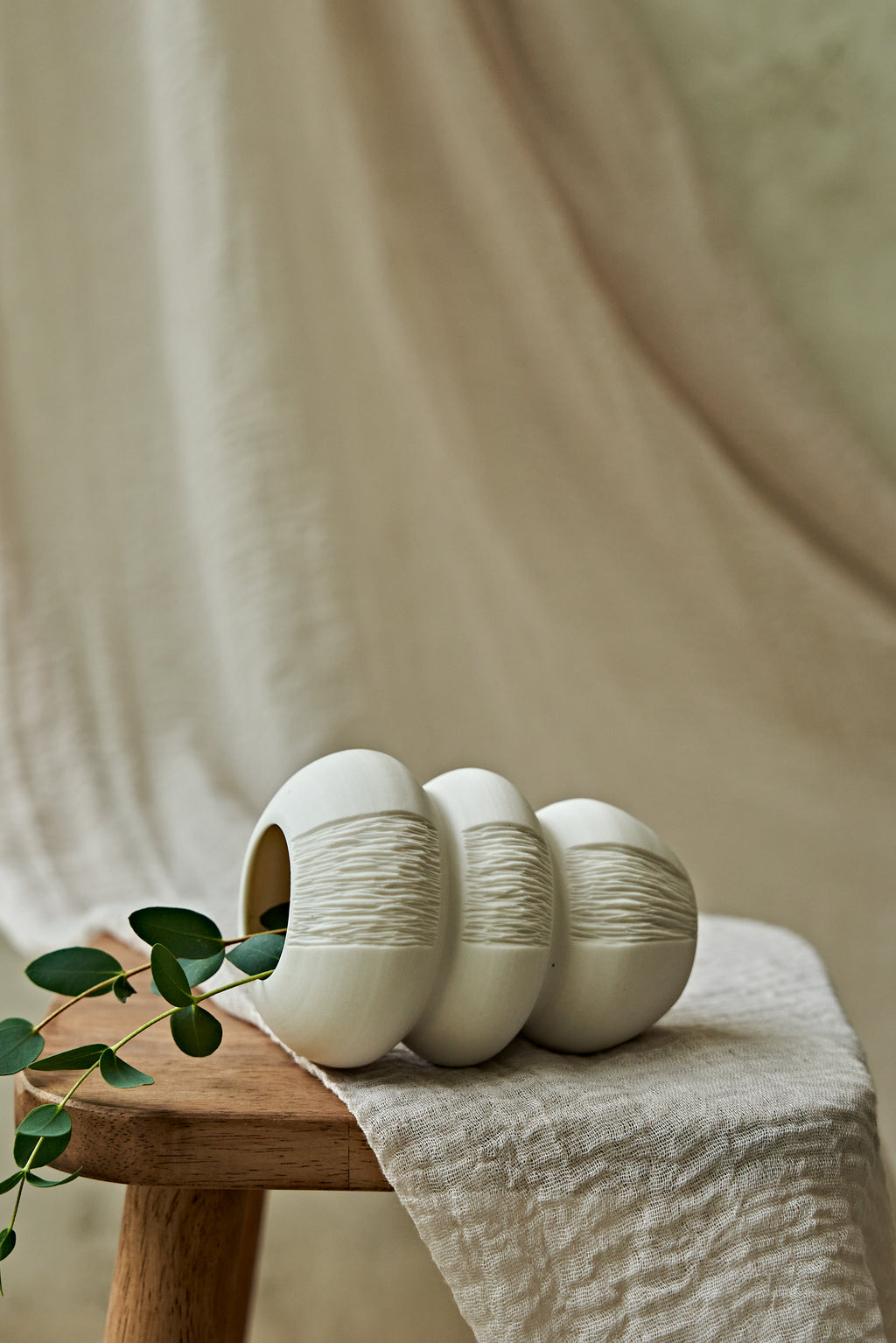 Three small white ceramic vases with textured vertical ridges, arranged on a wooden stool draped with a beige cloth, with a green leafy stem in the foreground.