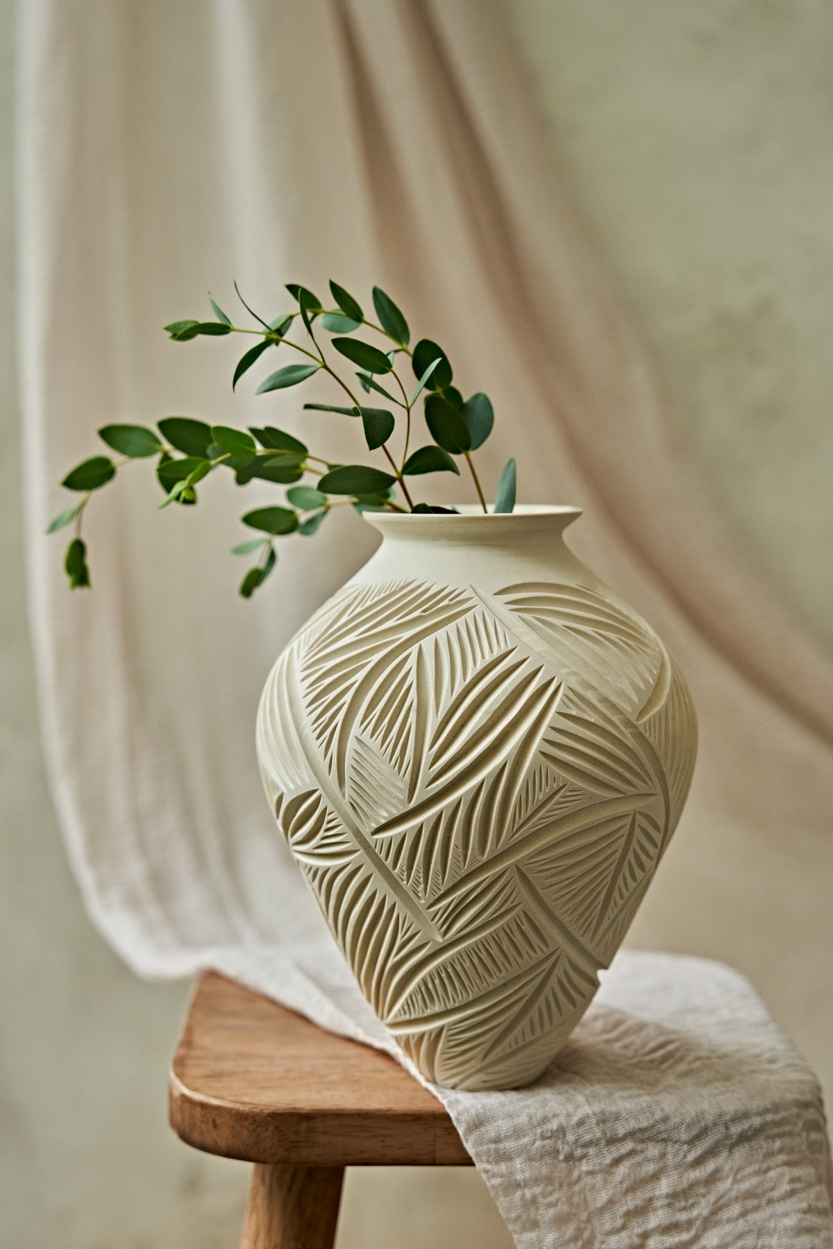 A cream-colored ceramic vase with a carved leaf pattern sits on a wooden stool draped with a white cloth, holding a green leafy branch against a soft beige fabric backdrop.