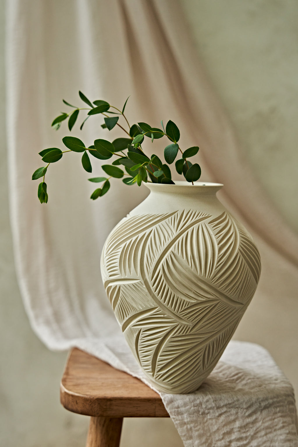 A cream-colored ceramic vase with a carved leaf pattern, holding a green leafy branch, styled on a wooden stool with draped fabric in the background.