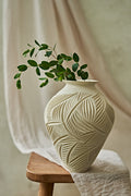 A cream-colored ceramic vase with a carved leaf pattern, holding a green leafy branch, styled on a wooden stool with draped fabric in the background.