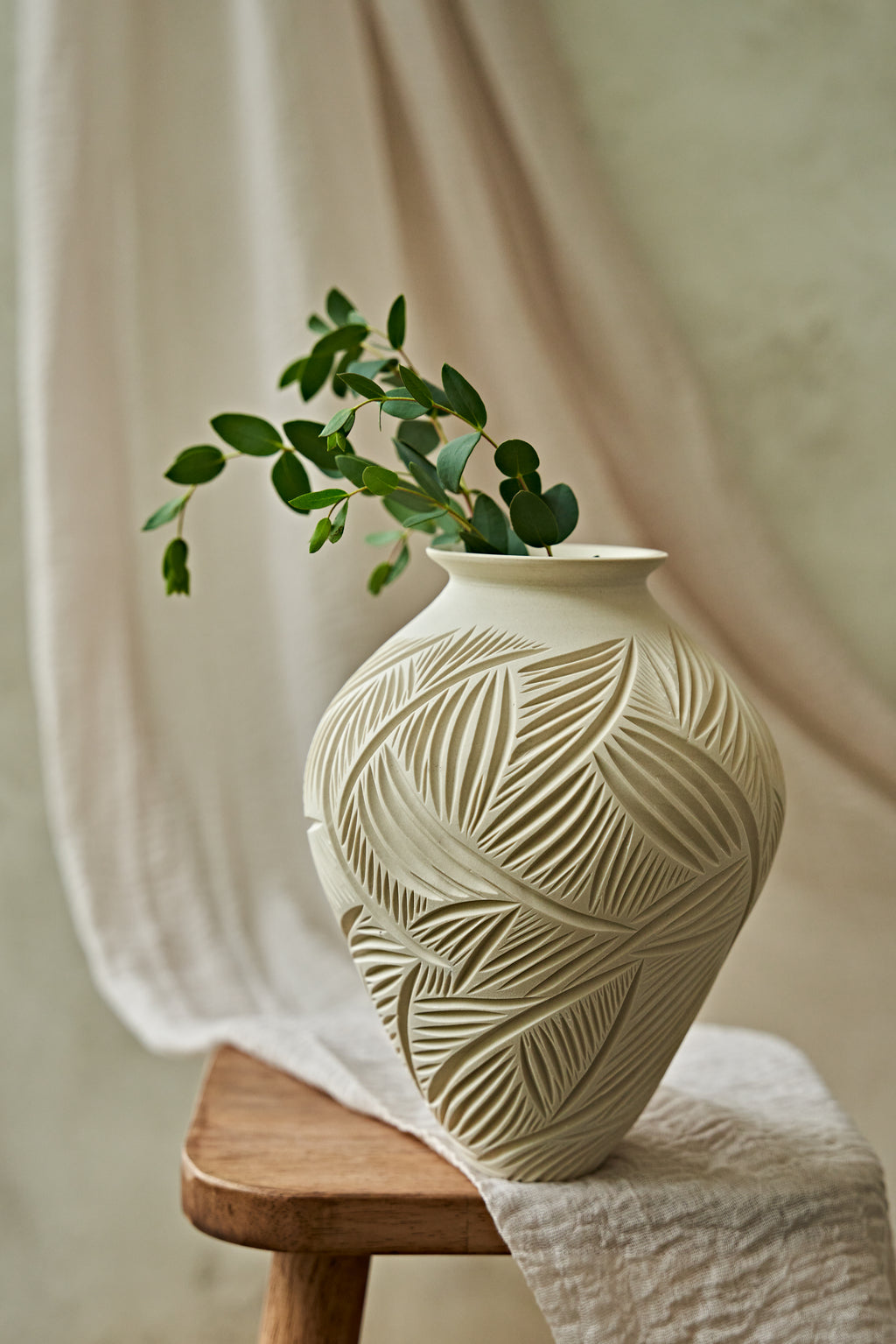 An off-white ceramic vase with a carved leaf pattern sits on a wooden stool draped with a white cloth, holding a green leafy branch against a soft beige background.