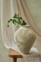 An off-white ceramic vase with a carved leaf pattern sits on a wooden stool draped with a white cloth, holding a green leafy branch against a soft beige background.