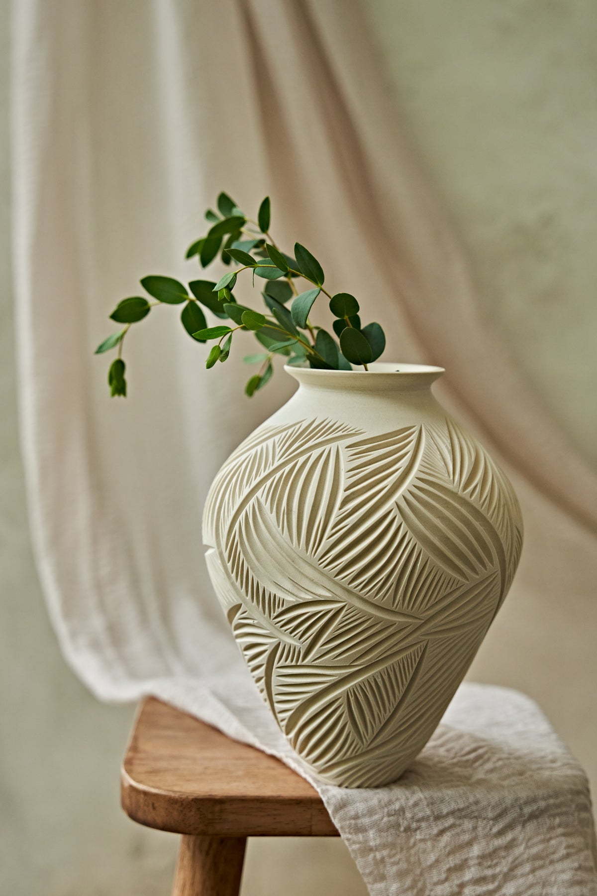 An off-white ceramic vase with a carved leaf pattern sits on a wooden stool draped with a white cloth, holding a green leafy branch against a soft beige background.