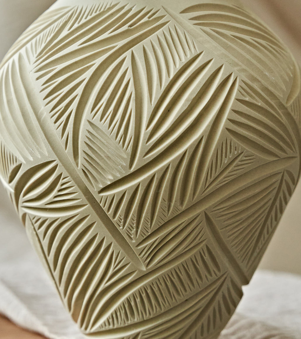 Close-up of a carved ceramic vase with a leaf pattern, in a warm beige color, resting on a white cloth.
