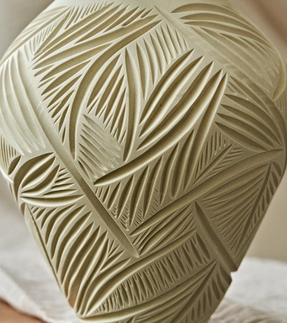 Close-up of a carved ceramic vase with a leaf pattern, in a warm beige color, resting on a white cloth.
