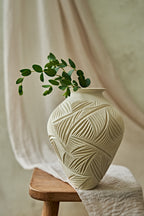 An off-white ceramic vase with a carved leaf pattern sits on a wooden stool draped with a cream fabric, holding a green leafy branch.
