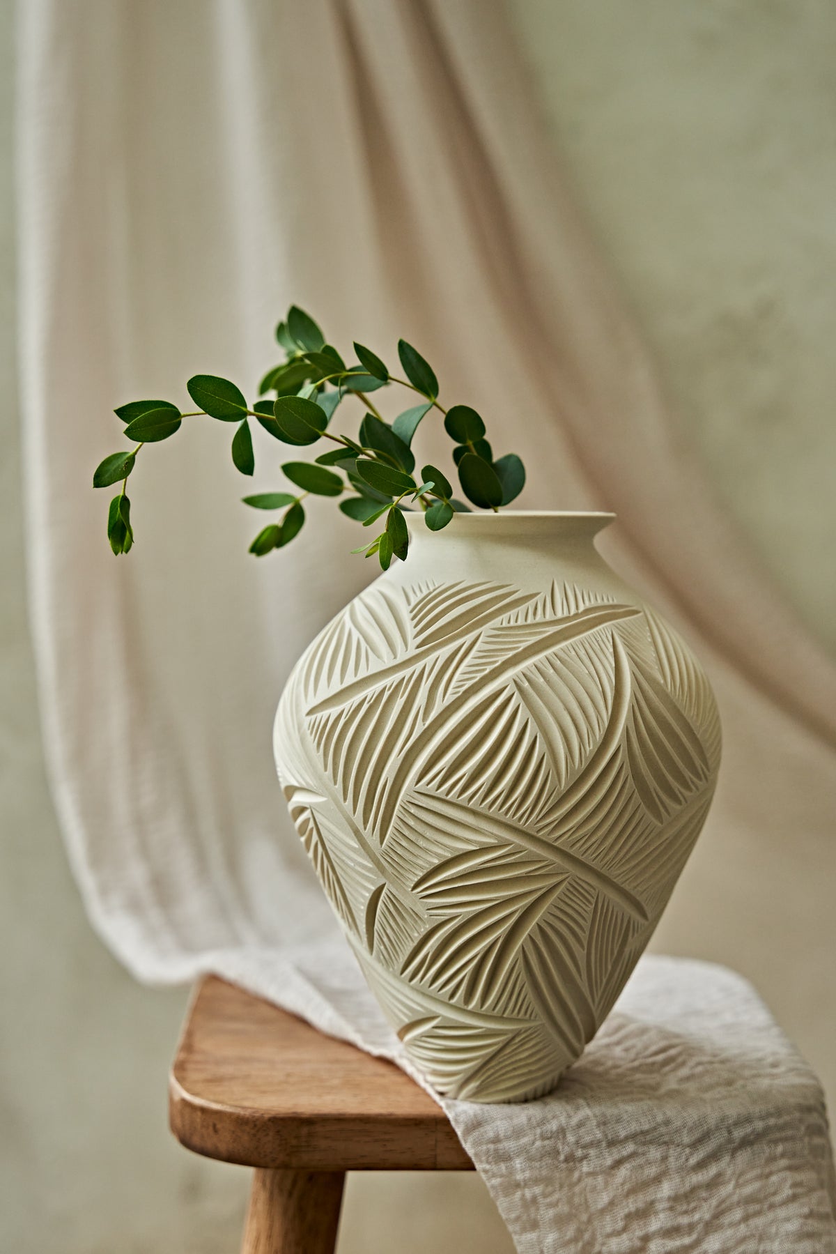 An off-white ceramic vase with a carved leaf pattern sits on a wooden stool draped with a cream fabric, holding a green leafy branch.