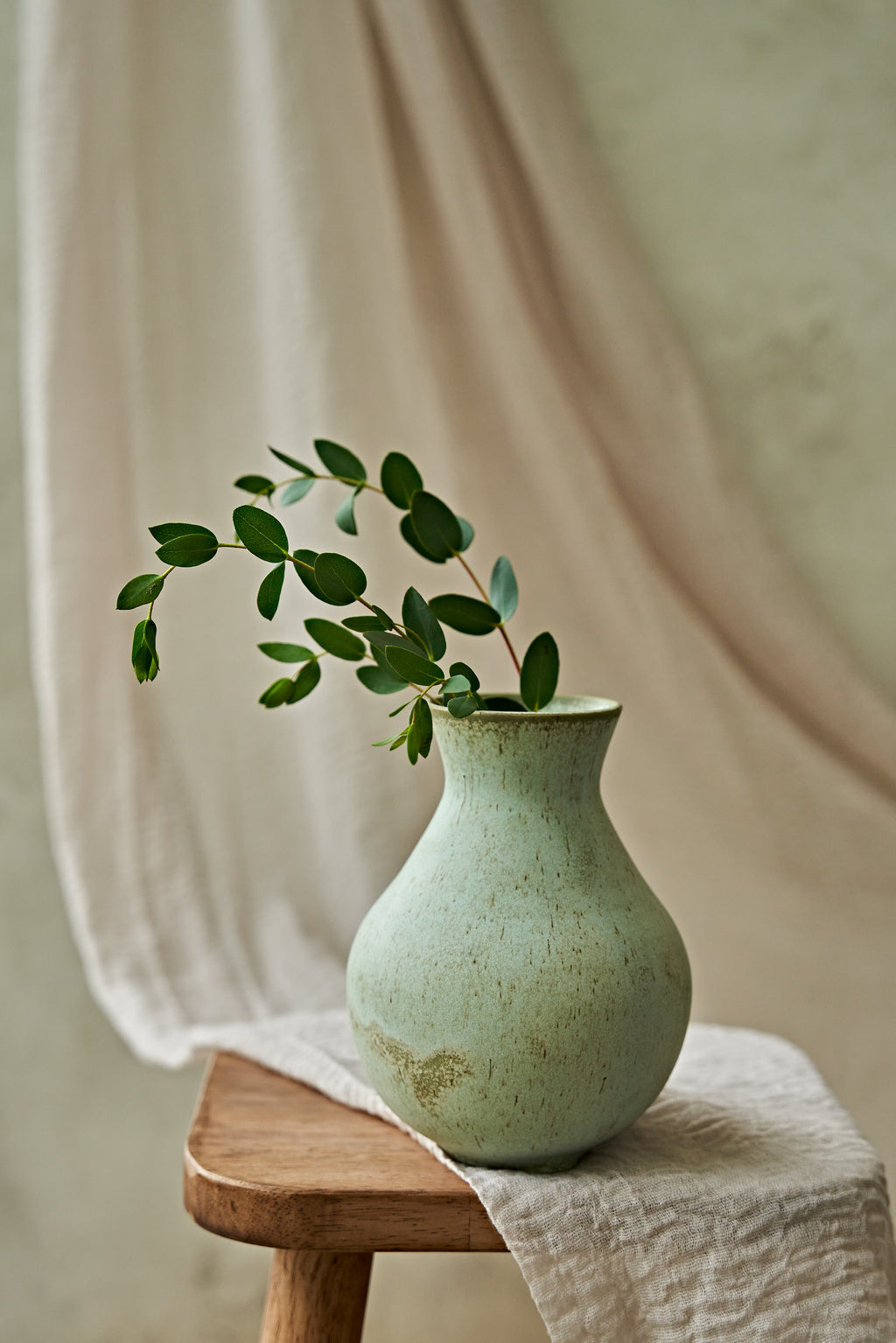 A light green speckled ceramic vase holding a green leafy branch, placed on a wooden stool draped with a beige cloth against a soft neutral background.