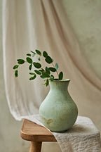 A light green speckled ceramic vase holding a green leafy branch, placed on a wooden stool draped with a beige cloth against a soft neutral background.