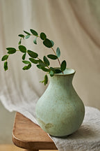 A small ceramic vase with a textured light green glaze sits on a wooden stool draped with a white cloth, holding a green leafy branch against a soft beige fabric backdrop.