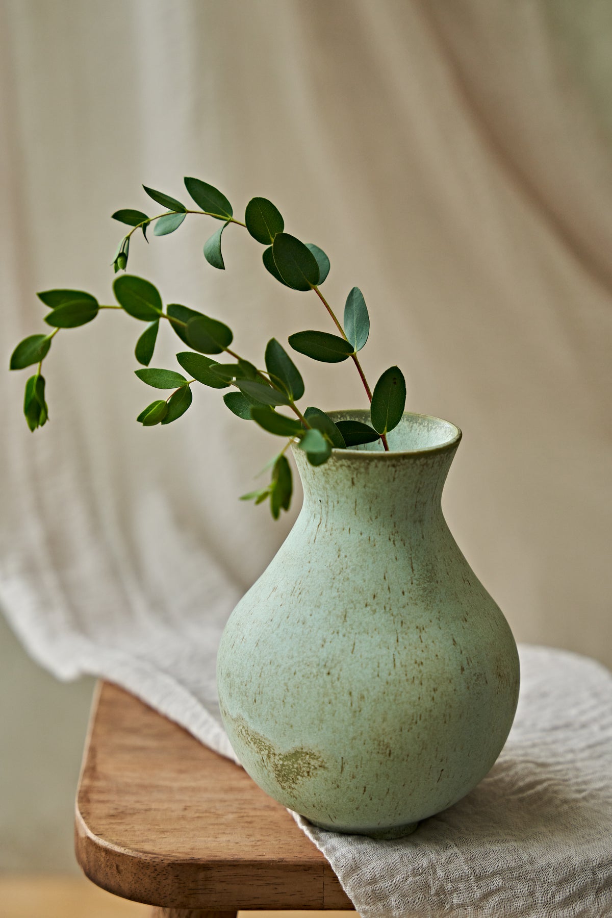 A small ceramic vase with a textured light green glaze sits on a wooden stool draped with a white cloth, holding a green leafy branch against a soft beige fabric backdrop.