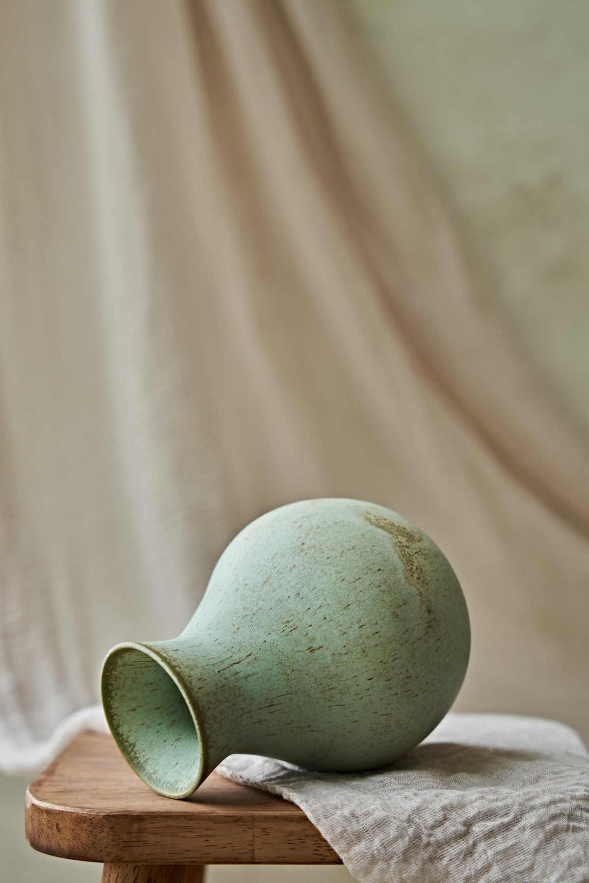 A small ceramic vase with a rounded bulbous body and a flared opening, finished in a matte pale green glaze with subtle earthy speckling. It is placed on a wooden stool draped with a light fabric, against a soft beige draped backdrop.