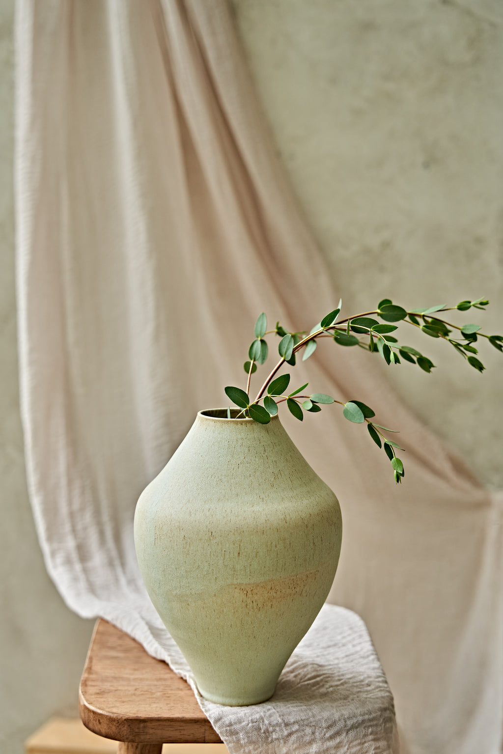 A light sage green ceramic vase with a textured finish, holding a delicate green leafy branch, styled on a wooden stool with draped beige fabric in the background.