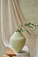 A light sage green ceramic vase with a textured finish, holding a delicate green leafy branch, styled on a wooden stool with draped beige fabric in the background.