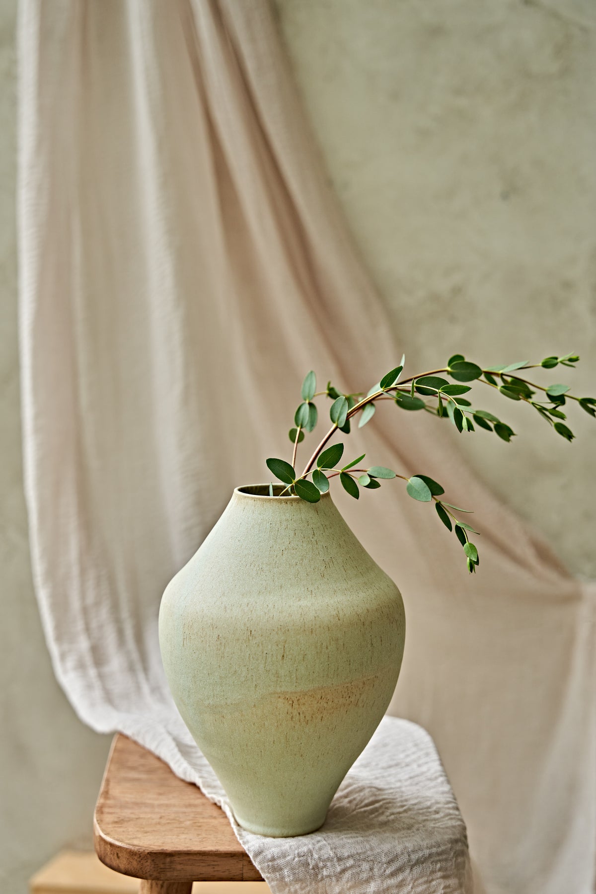 A light sage green ceramic vase with a textured finish, holding a delicate green leafy branch, styled on a wooden stool with draped beige fabric in the background.