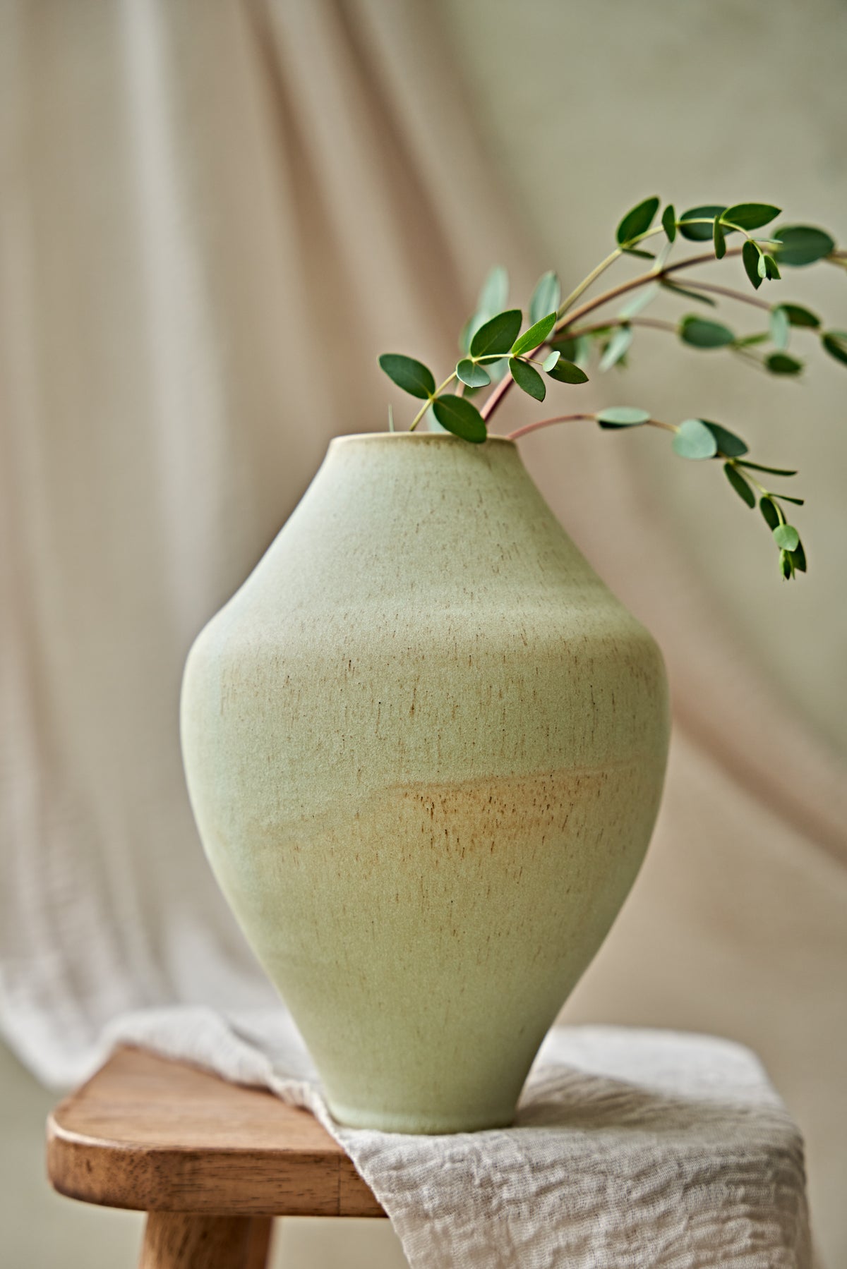 A light sage green ceramic vase with a textured glaze, styled on a wooden stool with a beige cloth and green eucalyptus branches, set against a soft beige draped fabric background.