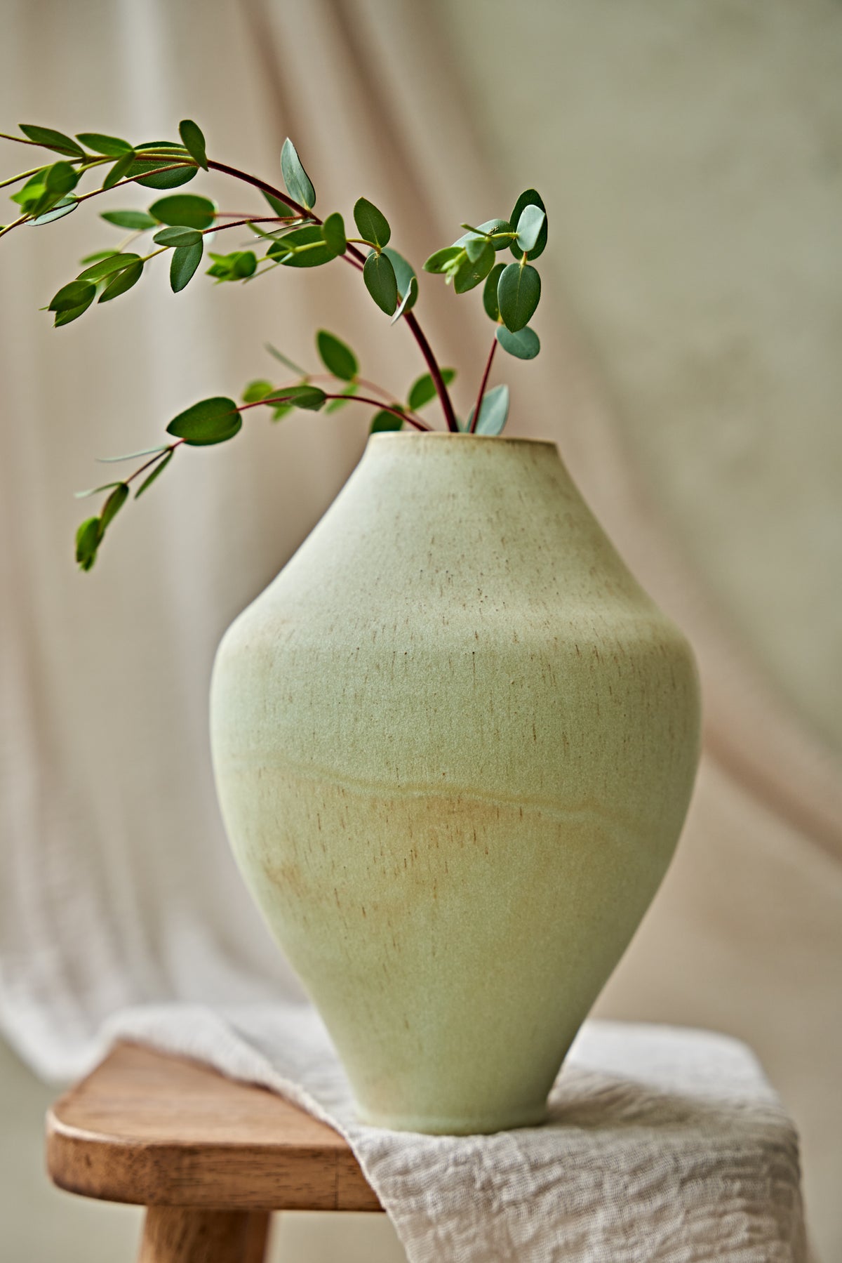 A light sage green ceramic vase with a textured matte finish, styled on a wooden stool with a draped linen cloth and green eucalyptus branches inside.