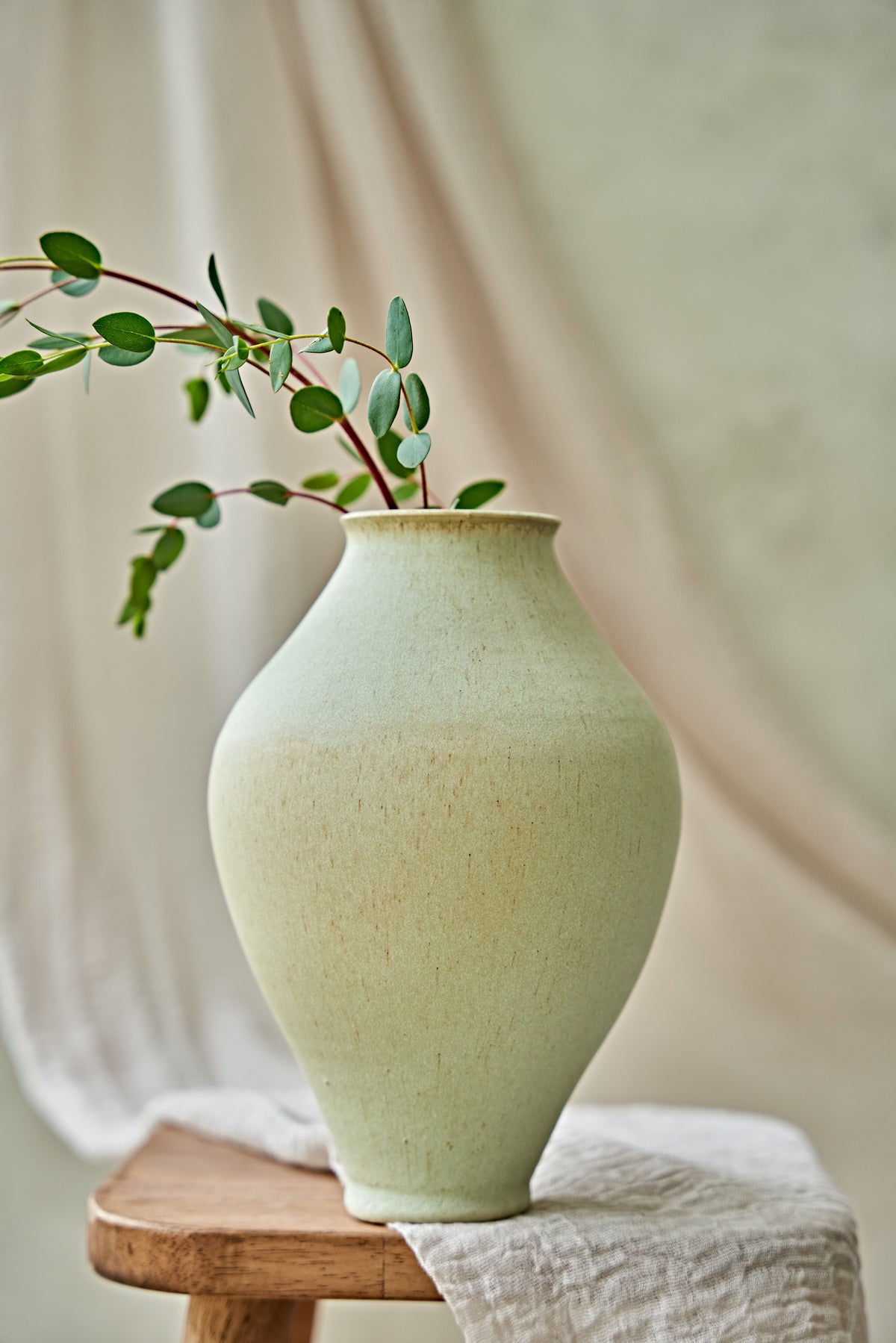 A light sage green ceramic vase with a textured matte finish, holding a delicate green eucalyptus sprig, resting on a wooden stool draped with a white linen cloth against a soft beige fabric backdrop.