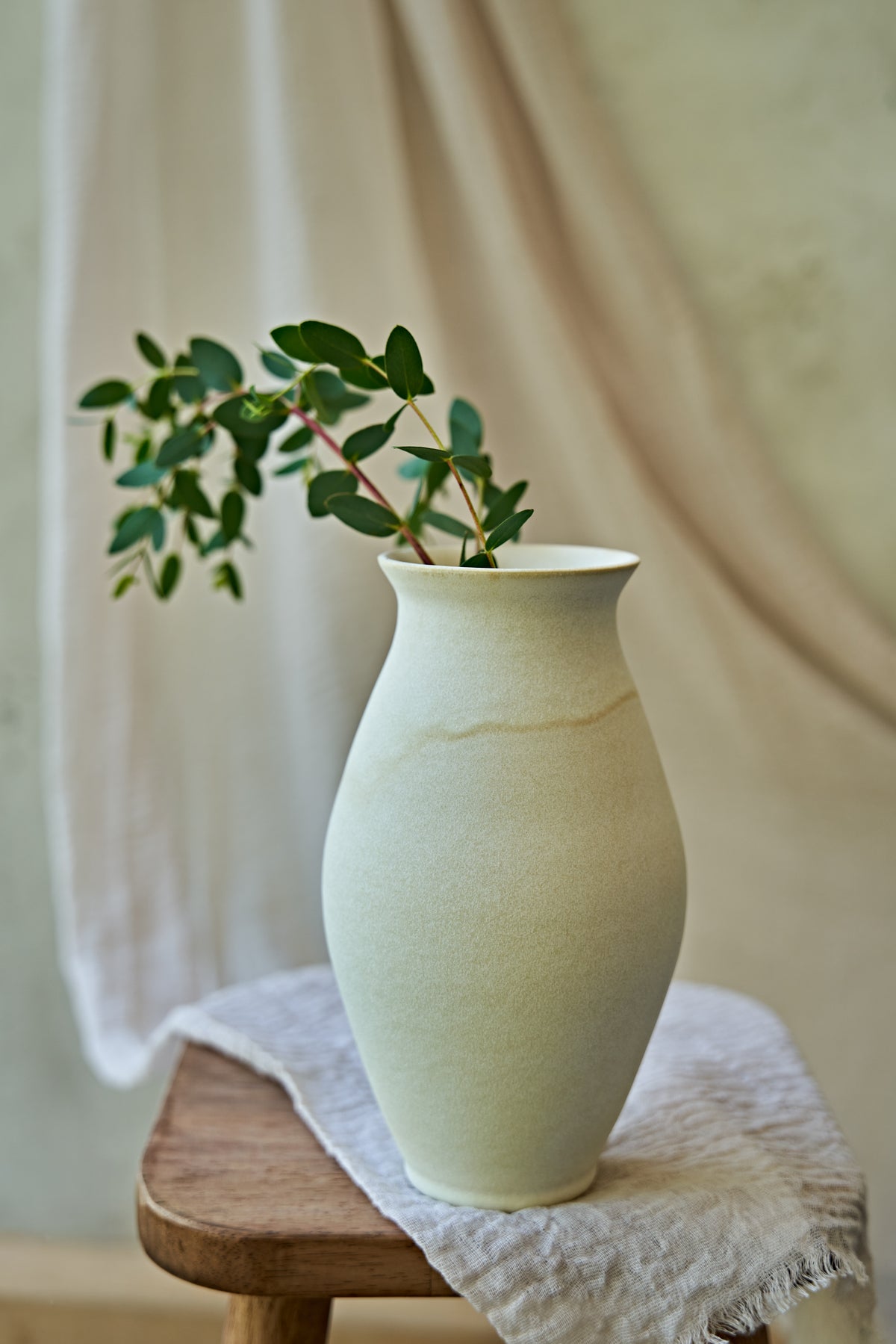 A large matte ceramic vase in a warm off-white or cream color, styled with green eucalyptus branches on a small wooden stool draped with a textured linen cloth against a soft beige background.