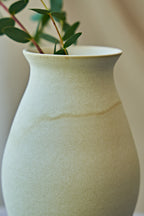 A close-up of a matte ceramic vase in a warm off-white or cream color, styled with green eucalyptus branches on a small wooden stool draped with a textured linen cloth against a soft beige background.