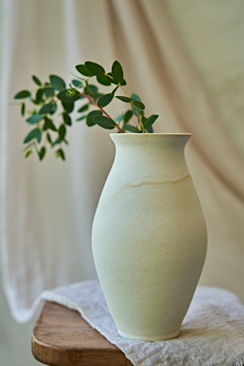 A large matte ceramic vase in a warm off-white or cream color, styled with green eucalyptus branches on a small wooden stool draped with a textured linen cloth against a soft beige background.
