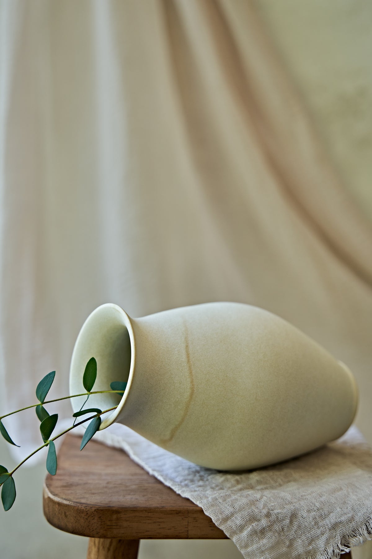 A large matte ceramic vase in a warm off-white or cream color, lying on its side and styled with green eucalyptus branches on a small wooden stool draped with a textured linen cloth against a soft beige background.
