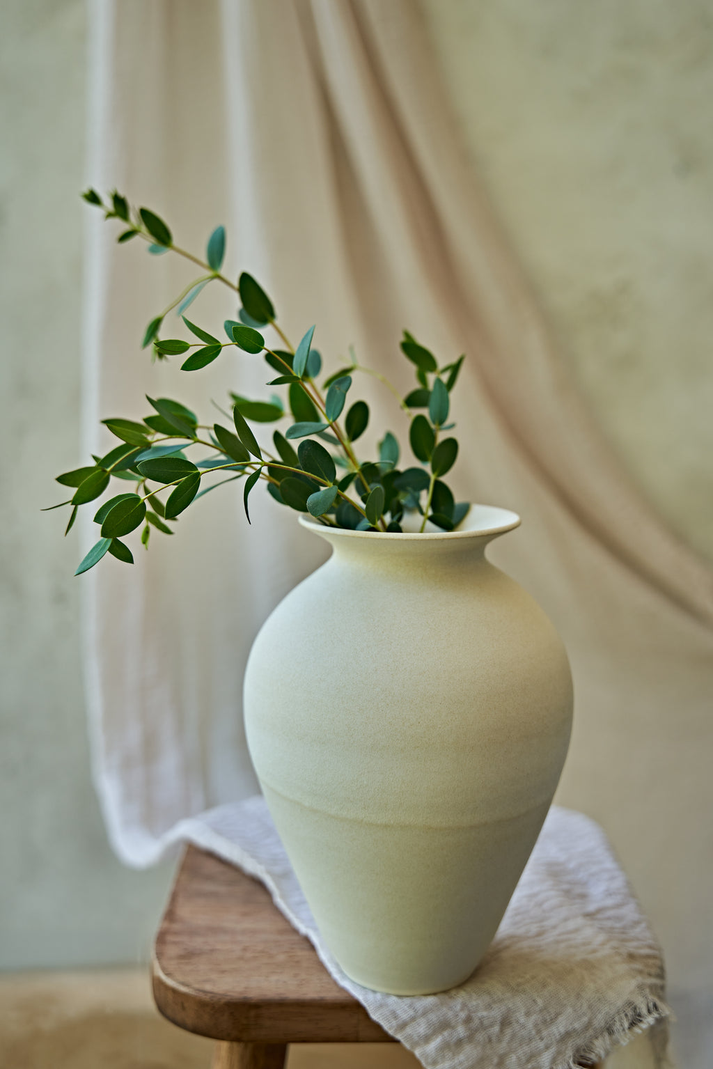 A large matte ceramic vase in a warm off-white or cream color, styled with green eucalyptus branches on a small wooden stool draped with a textured linen cloth against a soft beige background.