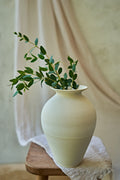 A large matte ceramic vase in a warm off-white or cream color, styled with green eucalyptus branches on a small wooden stool draped with a textured linen cloth against a soft beige background.